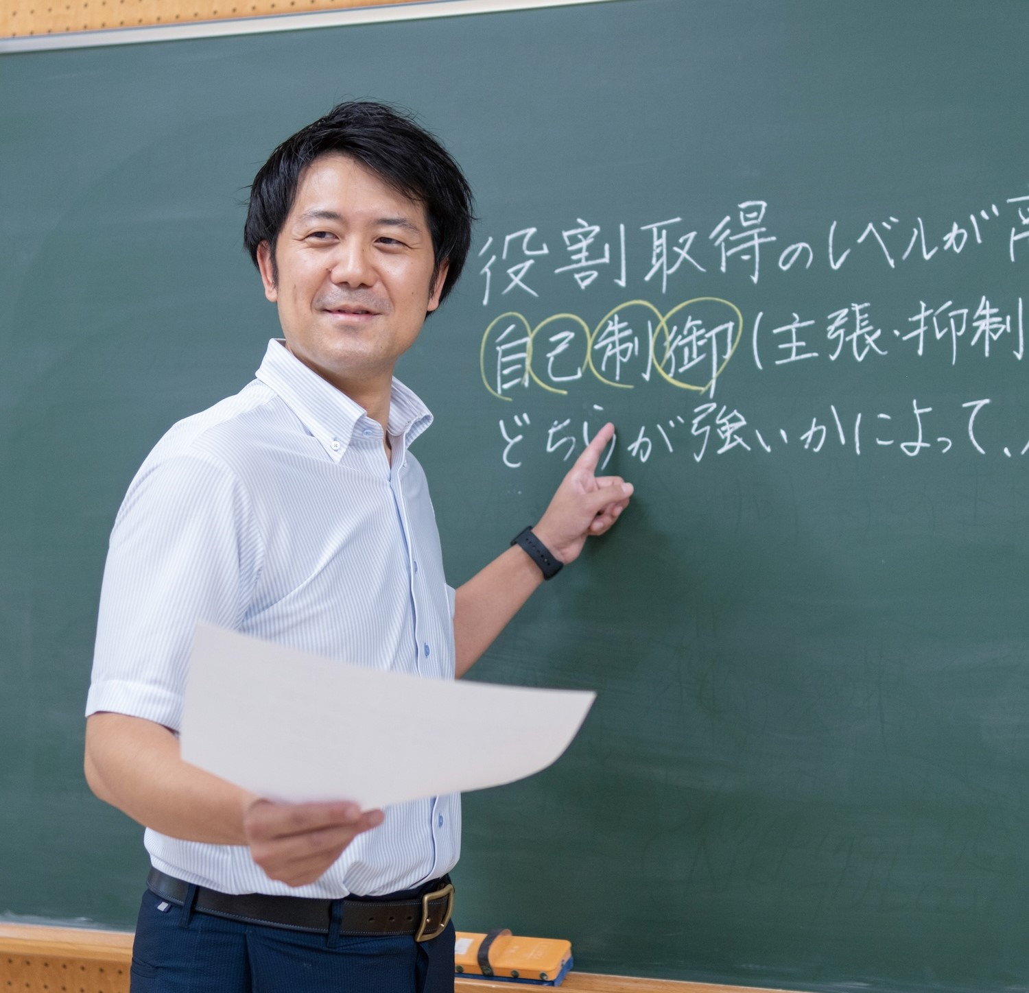 人間形成コース修了生の男性が、黒板に書いた文字を指差しながら、片手にプリントを持って生徒に教えている様子
